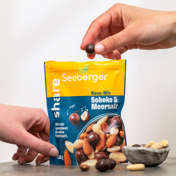 Verpackung des share-Mixes steht geöffnet da, eine Hand nimmt sich eine schokolierte Haselnuss aus der Packung