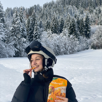 Person isst Trockenfrüchte, trägt Winterkleidung, steht fröhlich in schneebedeckter Waldlandschaft.