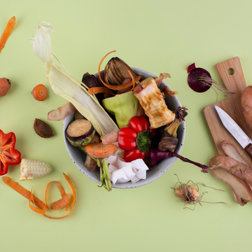 Schüssel mit Gemüseresten auf grünem Tisch, neben Schneidebrett mit Messer und Kartoffeln.