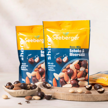 Zwei Packungen des share-Mixes stehen auf einem Holzbrett - davor liegen Nüsse und schokolierte Haselnusskerne