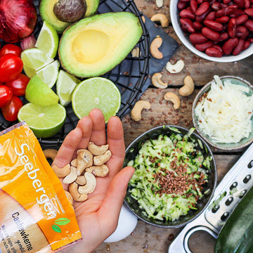 Hand hält Cashewkerne vor frischen Avocados, Limetten und anderen geschnittenen Zutaten auf Holztisch.