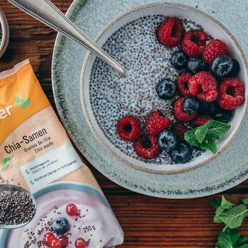 Schale Chia-Pudding mit Himbeeren und Blaubeeren, daneben eine Packung Chia-Samen, Holztisch.