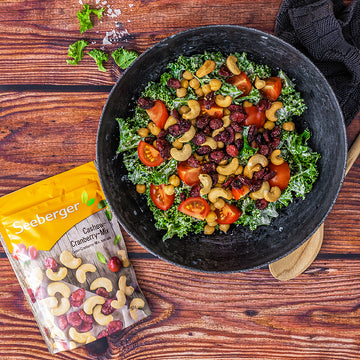 Salat mit Cashewkernen, Cranberrys und Tomaten liegt in schwarzer Schüssel auf Holztisch.
